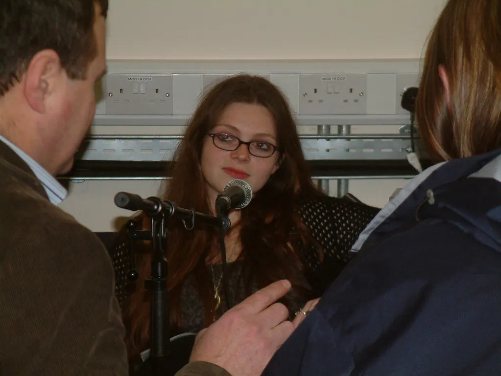 Twee mensen in gesprek aan een tafel in een podcaststudio, met microfoons op standaards en koptelefoons